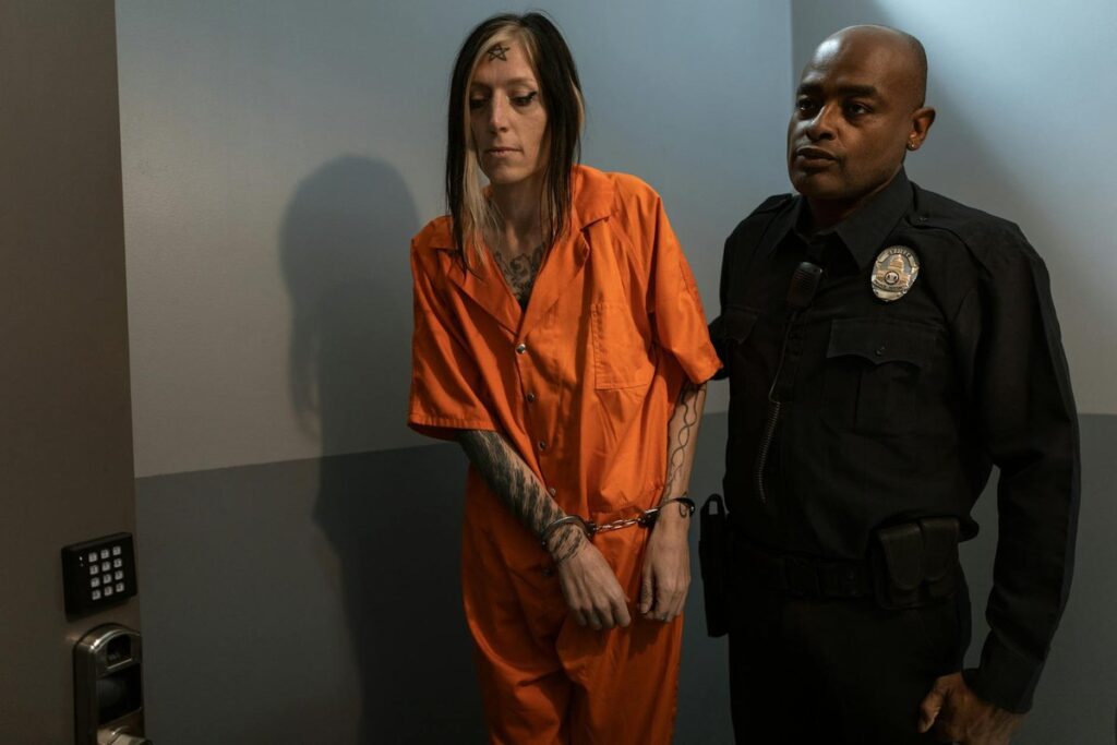 A handcuffed woman in an orange jumpsuit is escorted by a police officer indoors.