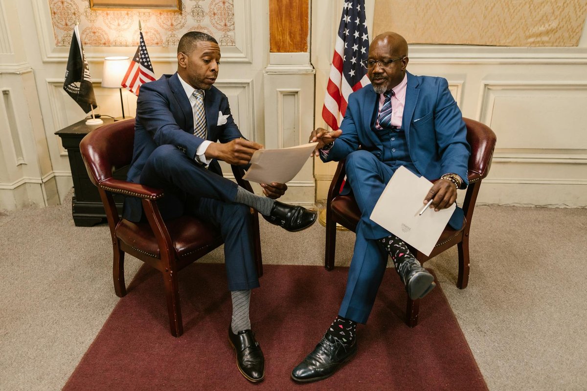 Two men in formal attire discussing documents in an office with flags and vintage decor.