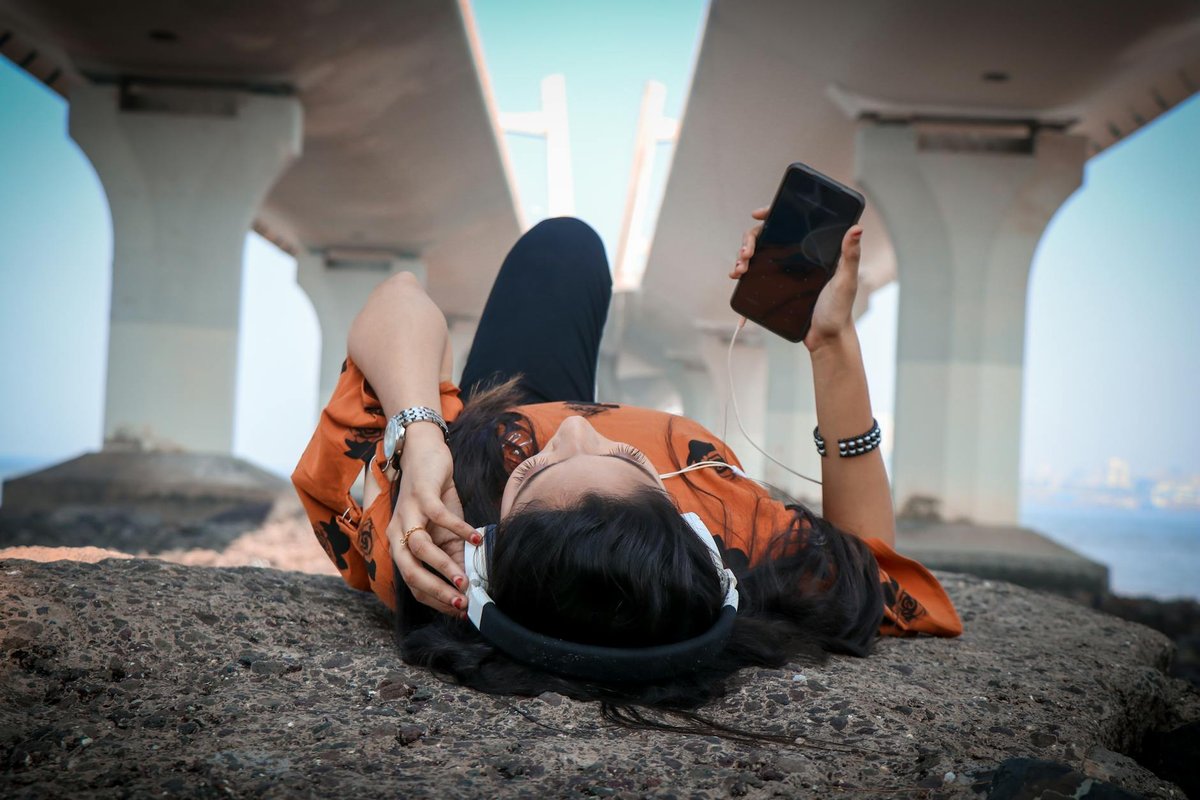 Woman lying under a bridge using headphones and phone, enjoying music outdoors.