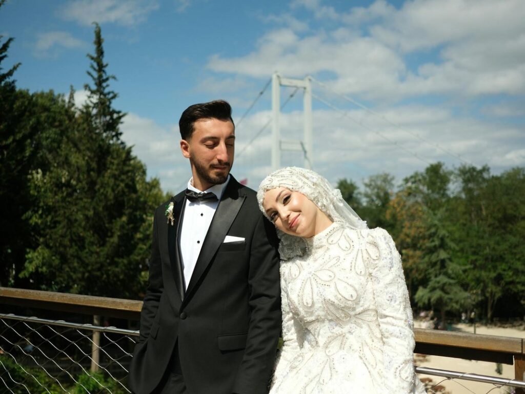 Bride and groom pose elegantly outdoors on a sunny day with a bridge backdrop.