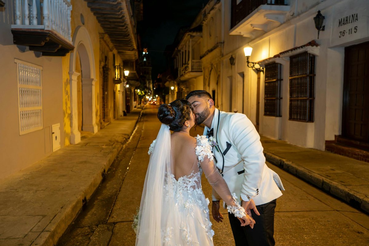 A couple shares a romantic kiss on a charming historic street at night, dressed in elegant wedding attire.