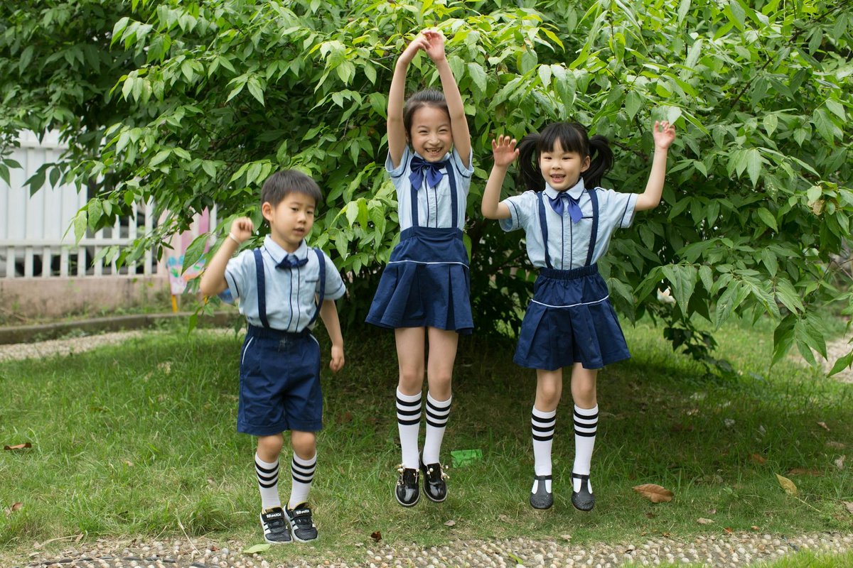 Three children in school uniforms jumping happily outdoors in a lush park setting.