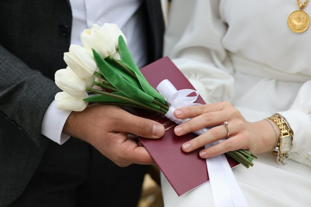 Close-up of a bride and groom holding white tulips and a red envelope during a wedding ceremony.