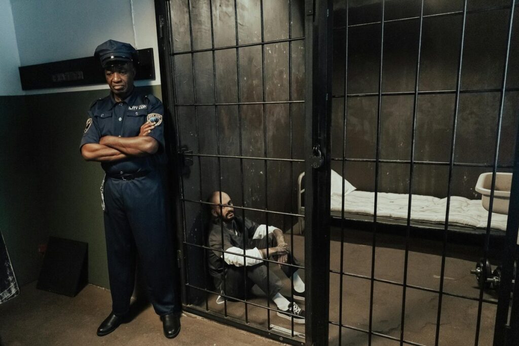 A police officer stands guard by a prison cell with a seated prisoner inside.