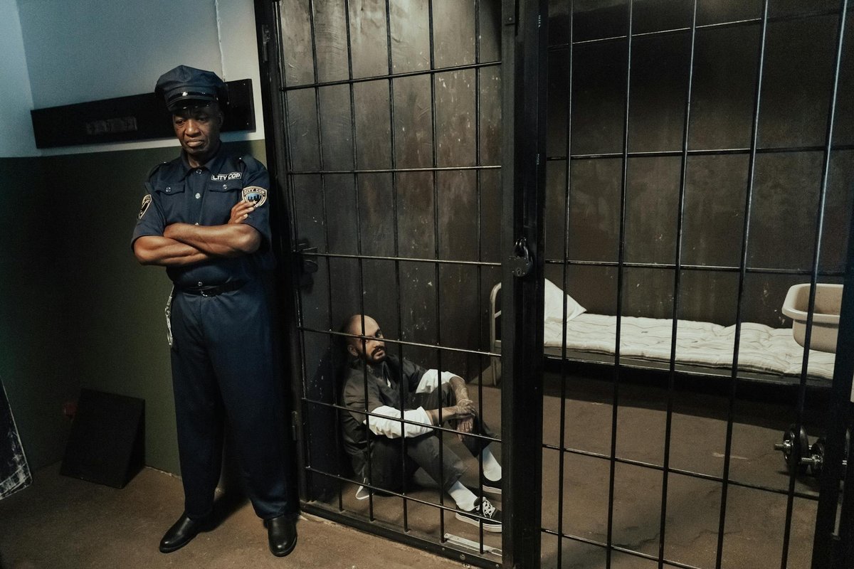 A police officer stands guard by a prison cell with a seated prisoner inside.