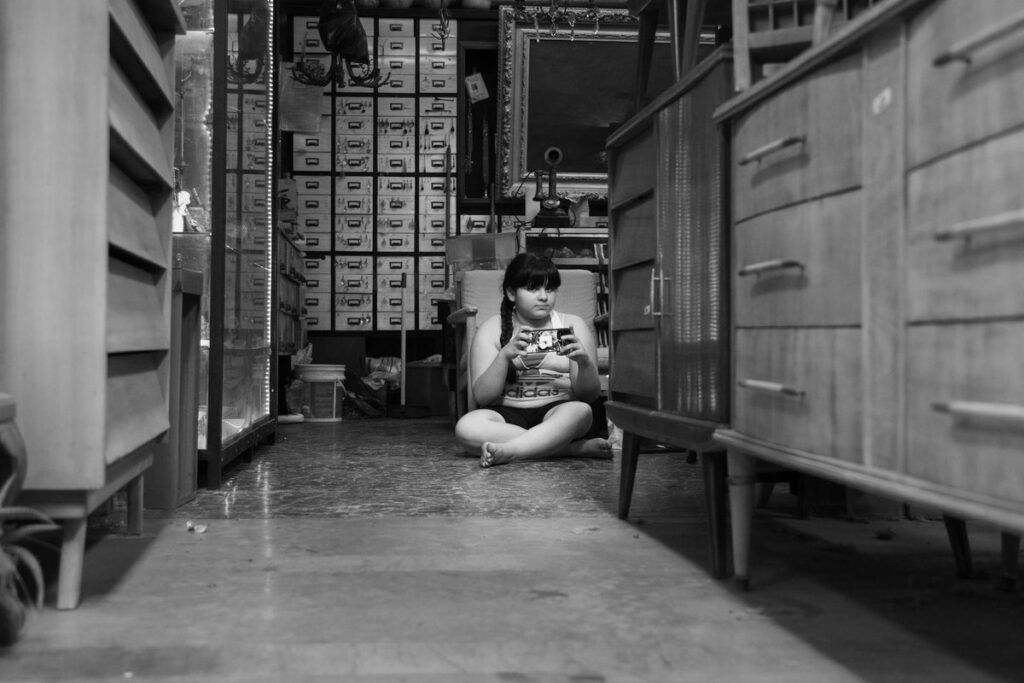 A child sits playing with a smartphone in a cluttered vintage shop filled with classic furniture.