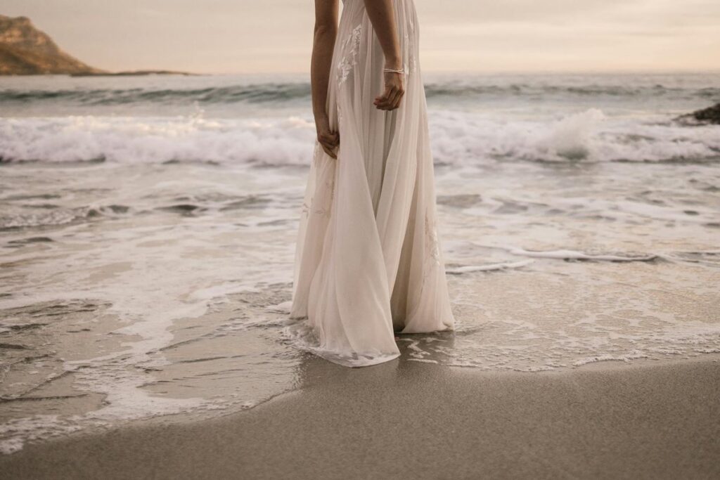 A bride in a wedding dress stands by the ocean at sunset, waves gently lapping at her feet.