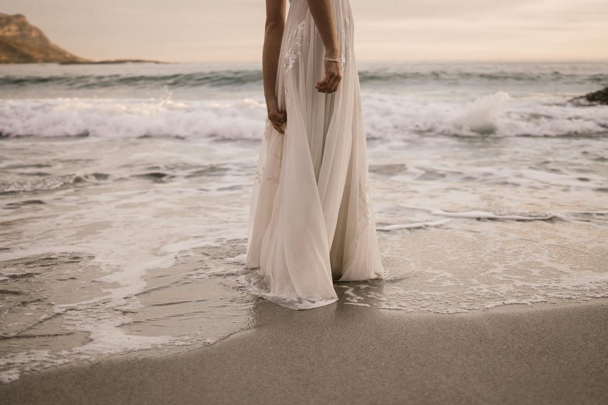 A bride in a wedding dress stands by the ocean at sunset, waves gently lapping at her feet.