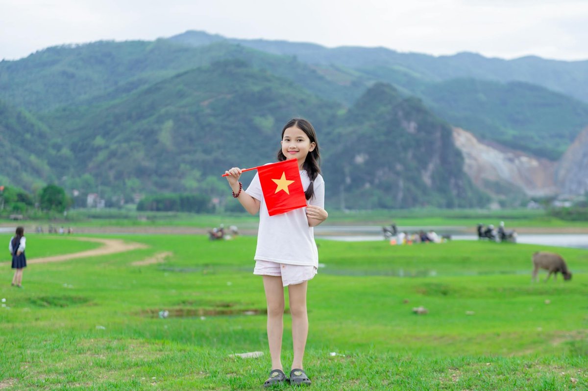 A young girl proudly holds a Vietnamese flag in a lush green field with mountains in the background.