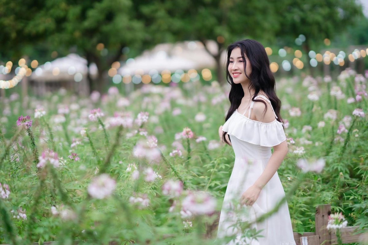 Asian woman in a white dress elegantly standing in a vibrant meadow with flowers.
