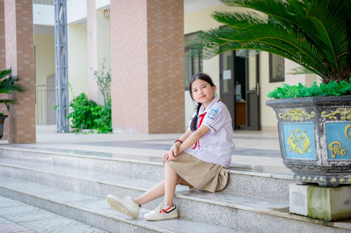 A young girl in a school uniform sits on steps outside a Vietnamese school building.