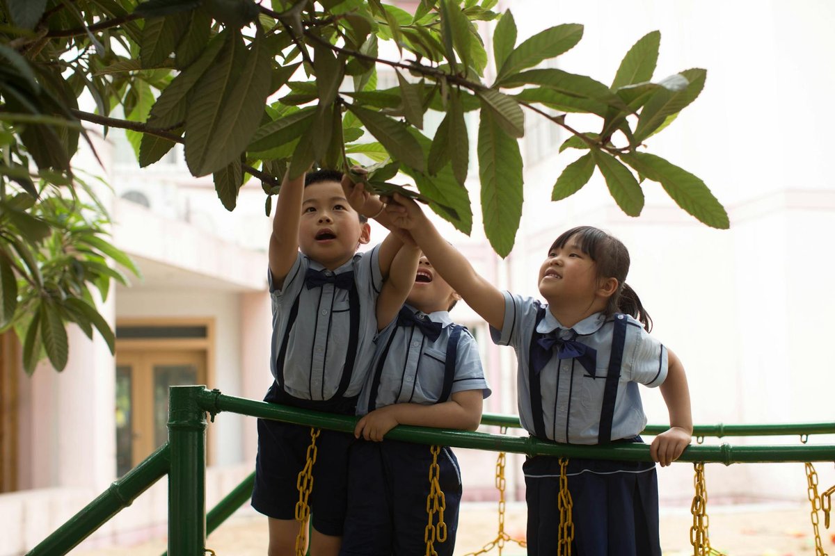 Three children in school uniforms reach for leaves on a playground.