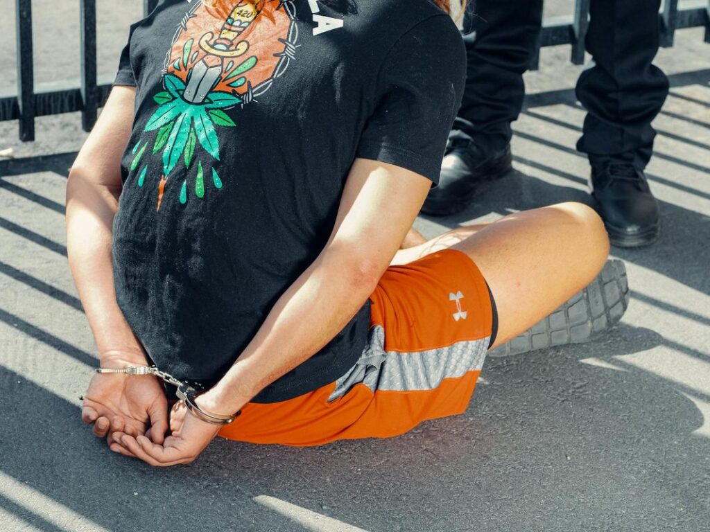 Person in handcuffs sitting on pavement, wearing athletic shorts and graphic t-shirt during daytime.