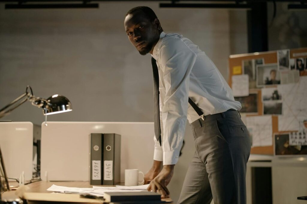 A detective stands at a desk, analyzing documents, in a well-lit office environment.