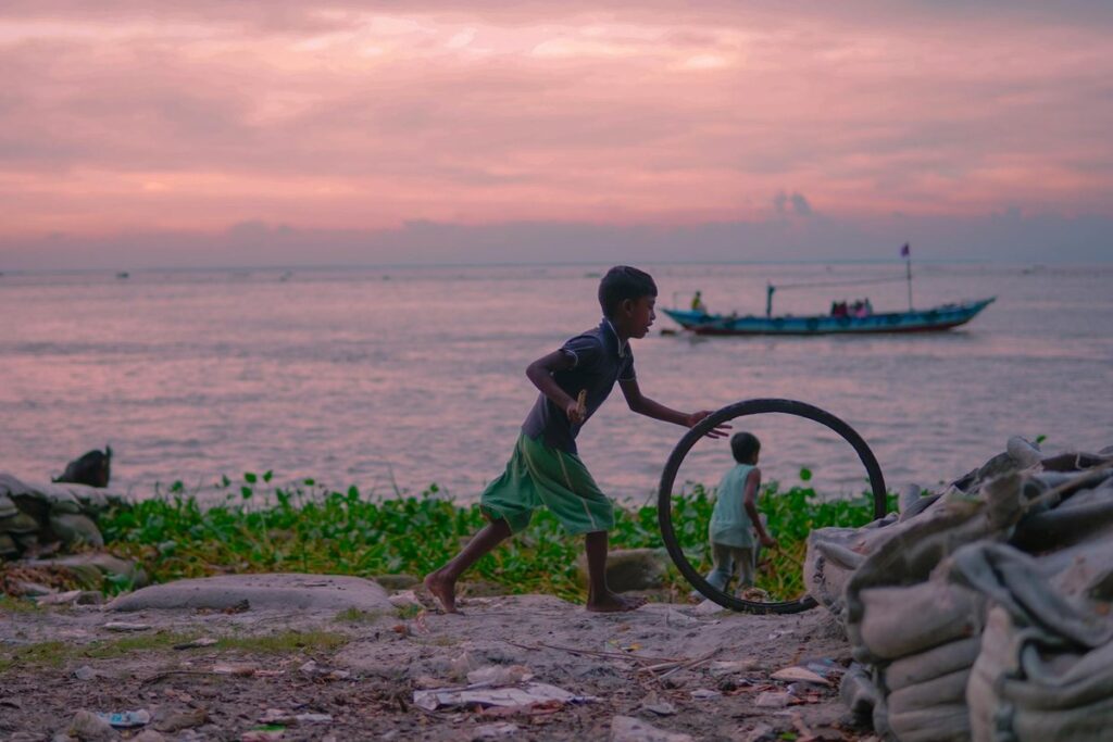 Two children playing with a tire by the riverbank during a beautiful sunset with a boat in the background.