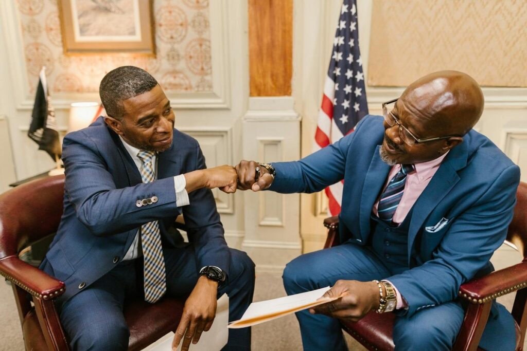 Two businessmen in suits engage in a friendly fist bump in a formal office environment.