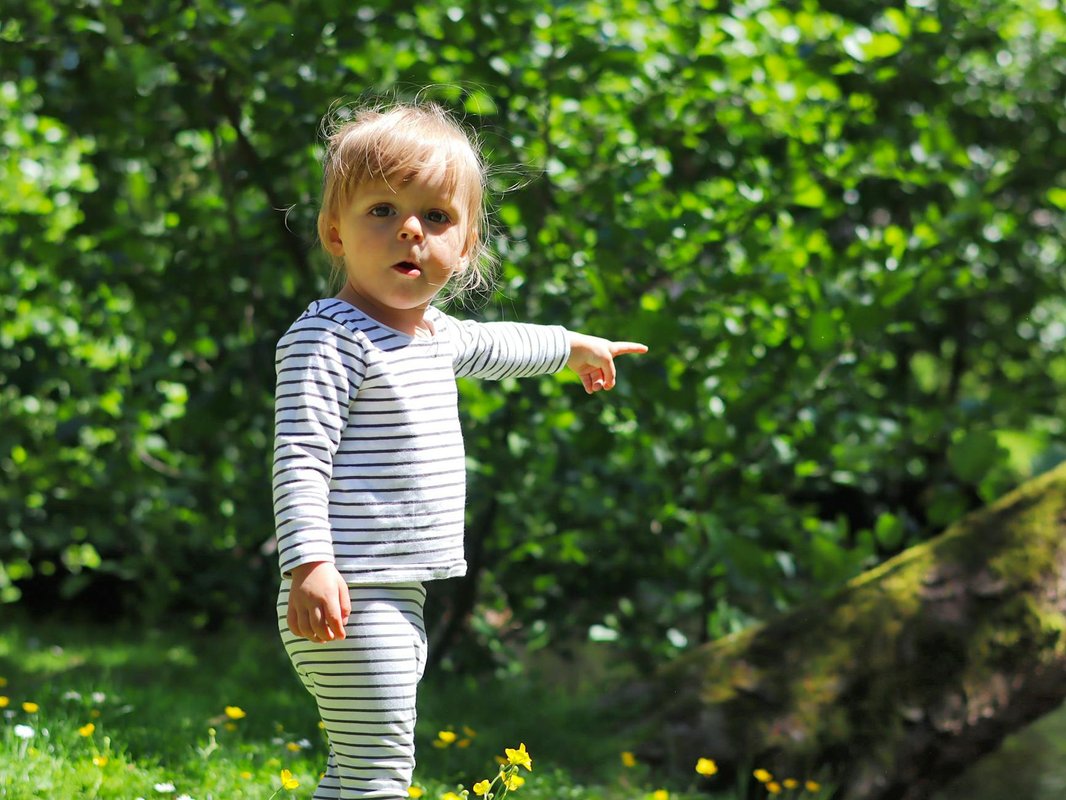 A toddler in striped outfit pointing forward in a sunlit park with dense greenery.
