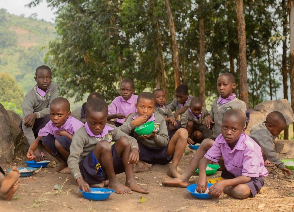Group of children enjoying a meal outdoors in a rural area, wearing school uniforms.