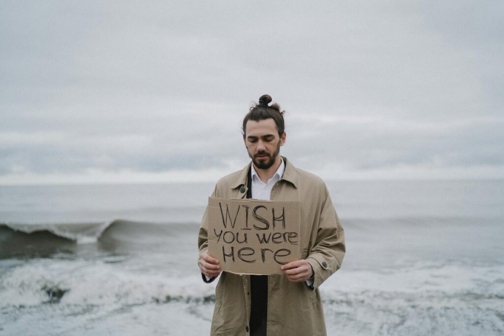 A man in a trench coat stands by the sea holding a 'Wish you were here' sign, expressing longing.