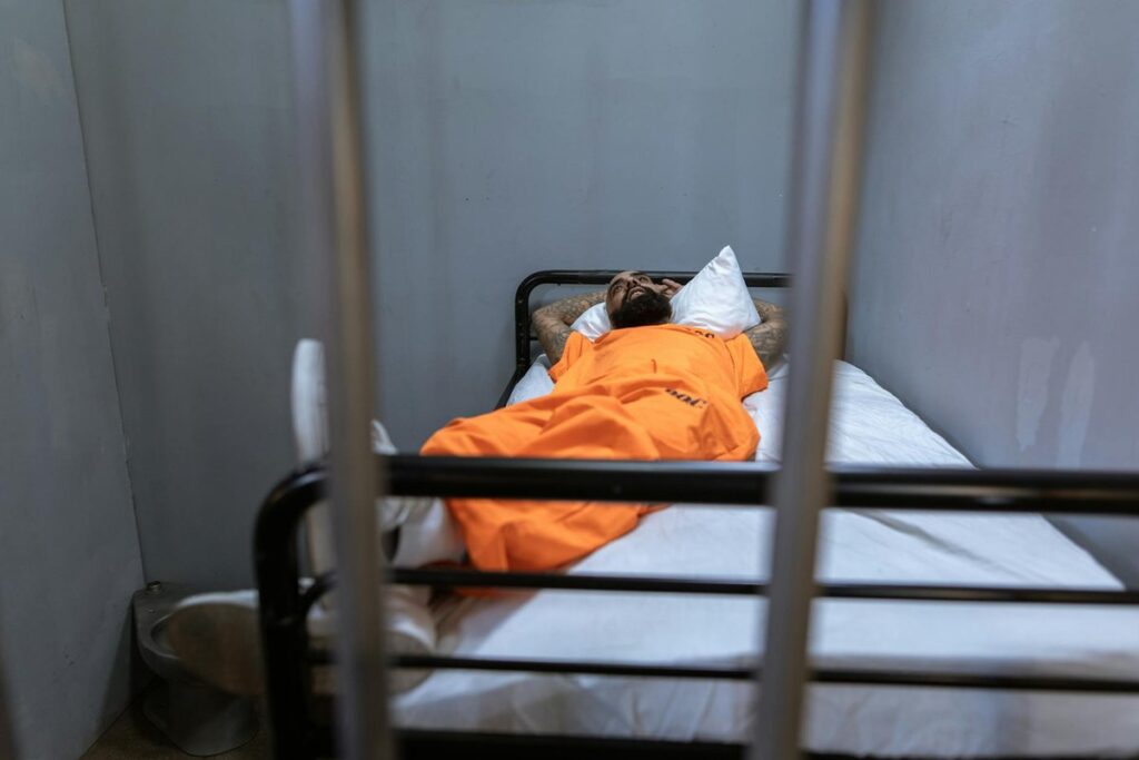 A prisoner in an orange jumpsuit lying on a bed inside a prison cell, viewed through metal bars.