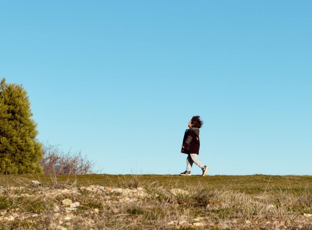 Little Boy Walking on Green Hill Against Blue Sky