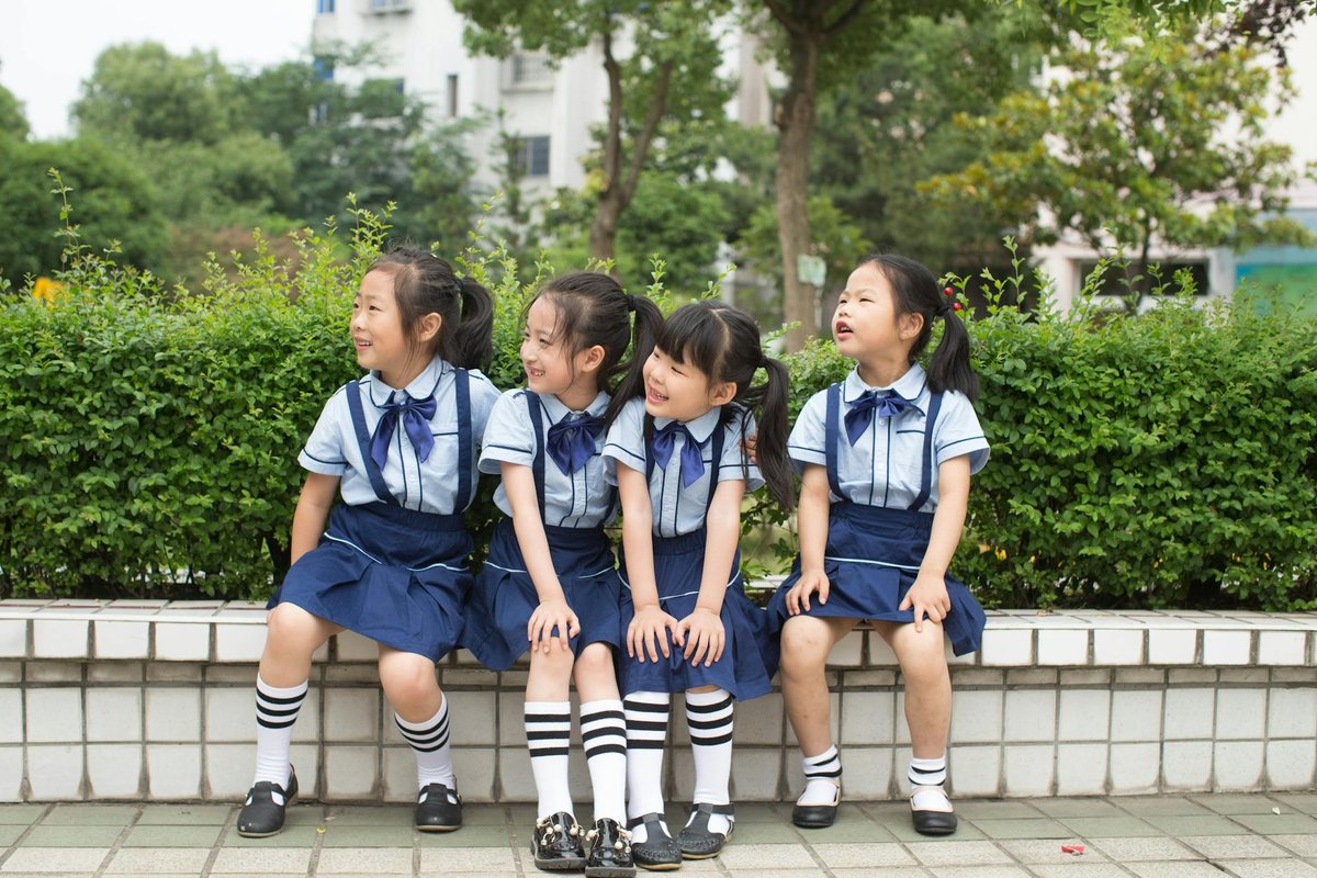 Four young girls in school uniforms sitting on a bench outdoors, smiling and looking to the side.