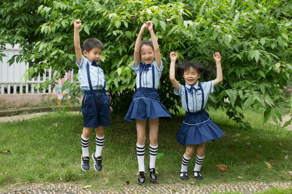 Three smiling children in school uniforms jumping joyfully outdoors in summer.