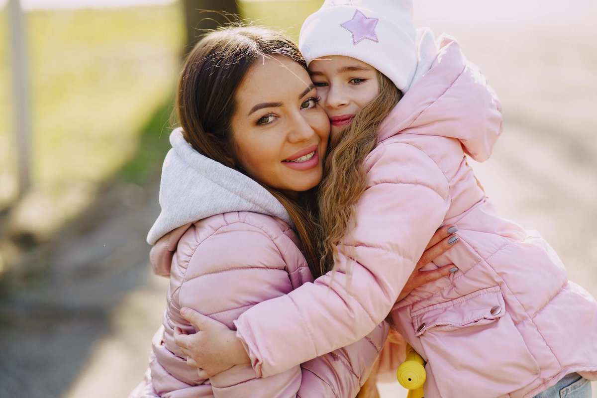 Joyful mother and daughter embracing in a sunny park during spring wearing pink jackets.