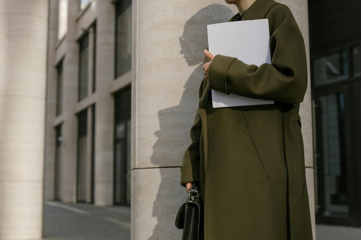 Woman in a green coat holding documents outside a modern office building.
