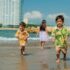 Three children playing happily on a sunny beach with the sea and buildings in the background.