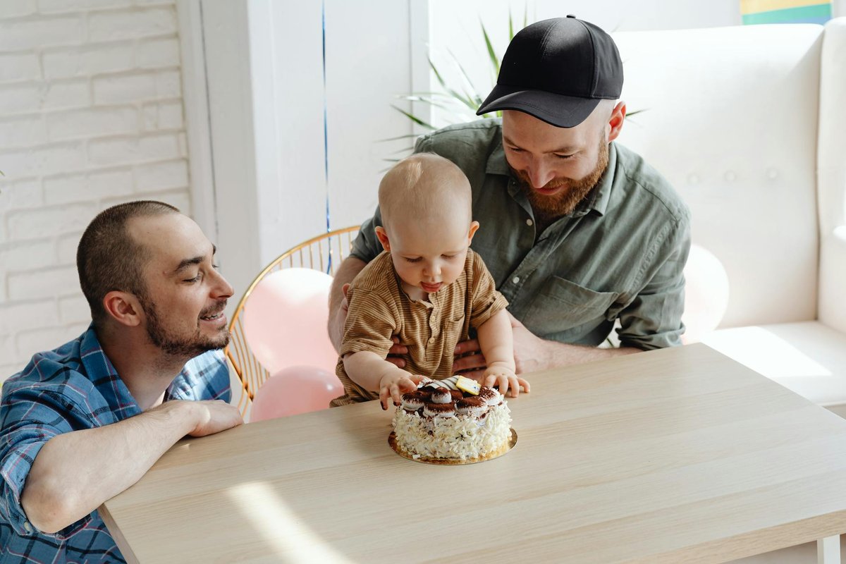Two fathers with their baby celebrating a birthday with a cake indoors.
