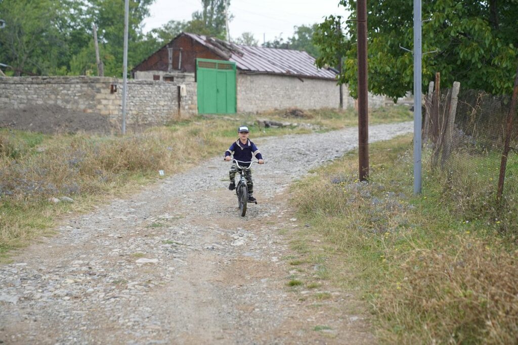 Young child riding a bicycle on a rural dirt road near village houses.