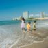 Three children play joyfully on a sunny beach with a city skyline in the background.