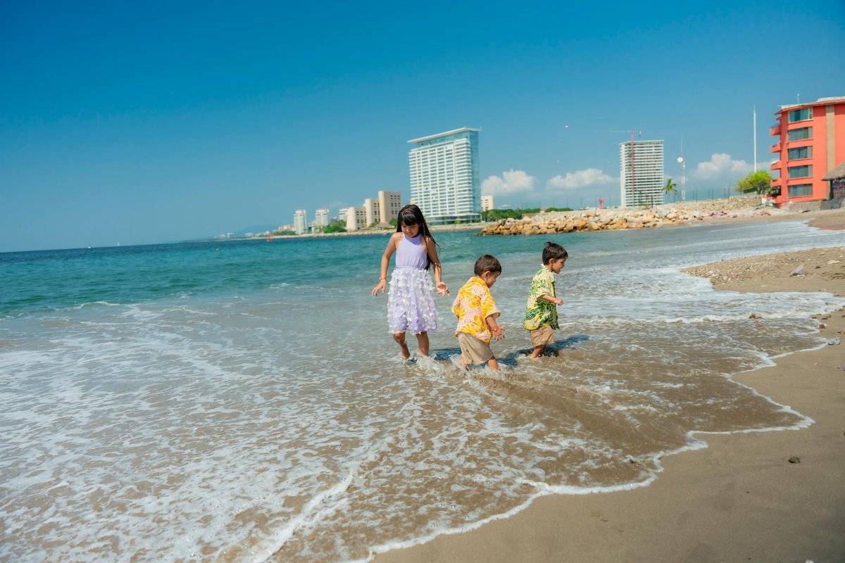 Three children play joyfully on a sunny beach with a city skyline in the background.