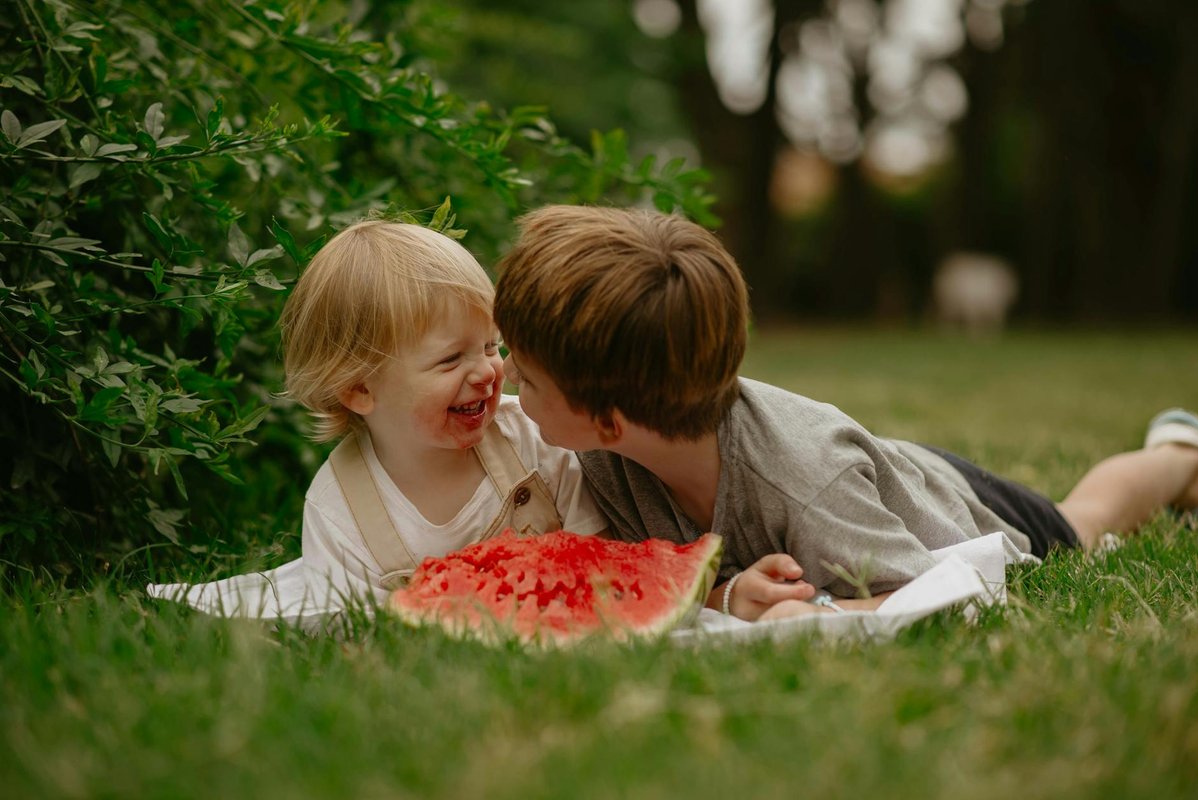 Two young children laughing and sharing a watermelon slice on a sunny summer day outdoors.