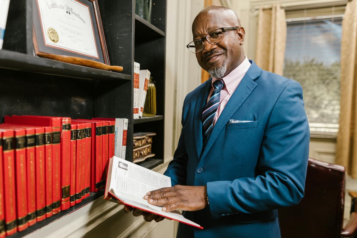 Professional lawyer in formal attire holding a book, smiling in an office setting surrounded by law books.