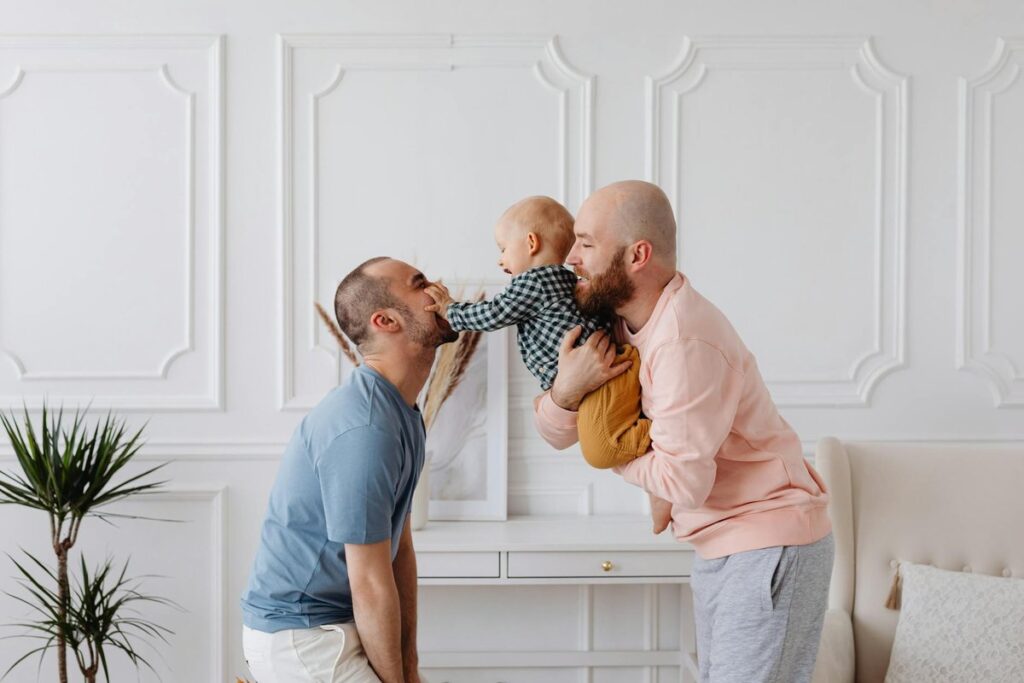 Two fathers joyfully interacting with their smiling toddler, capturing love and family ties indoors.