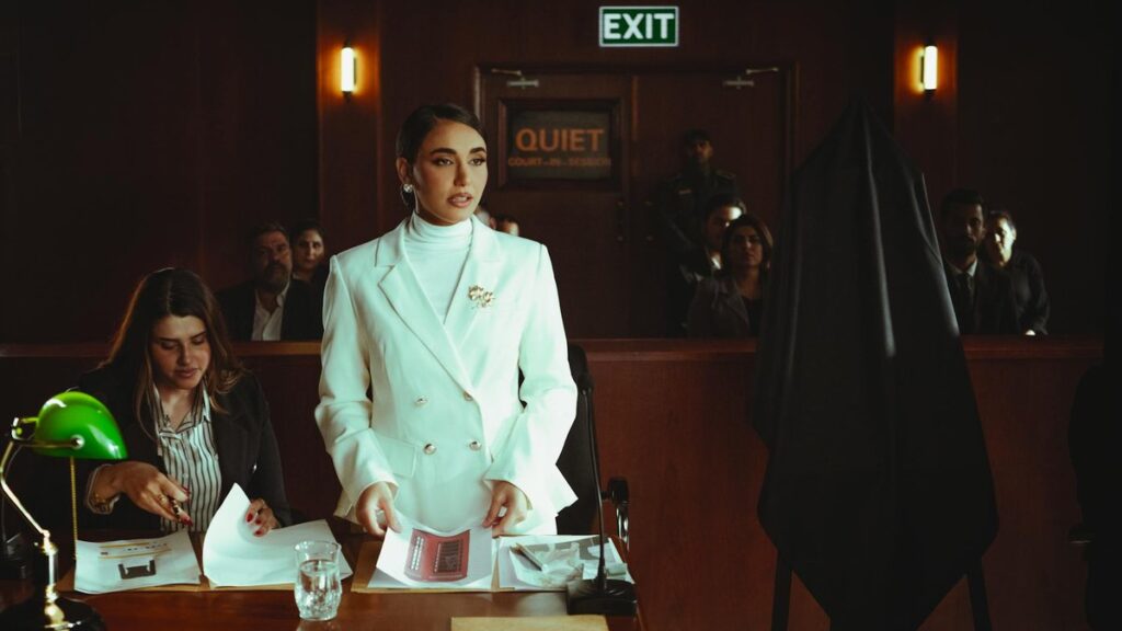 A lawyer in white suit confidently presents a case during a courtroom session with colleagues and audience.
