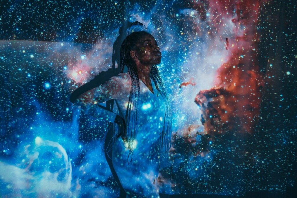 A woman with braided hair poses, surrounded by a cosmic projection, evoking a celestial theme.