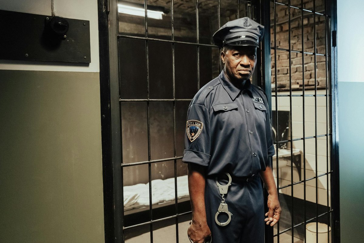 A prison guard in uniform standing in front of a cell inside a correctional facility.