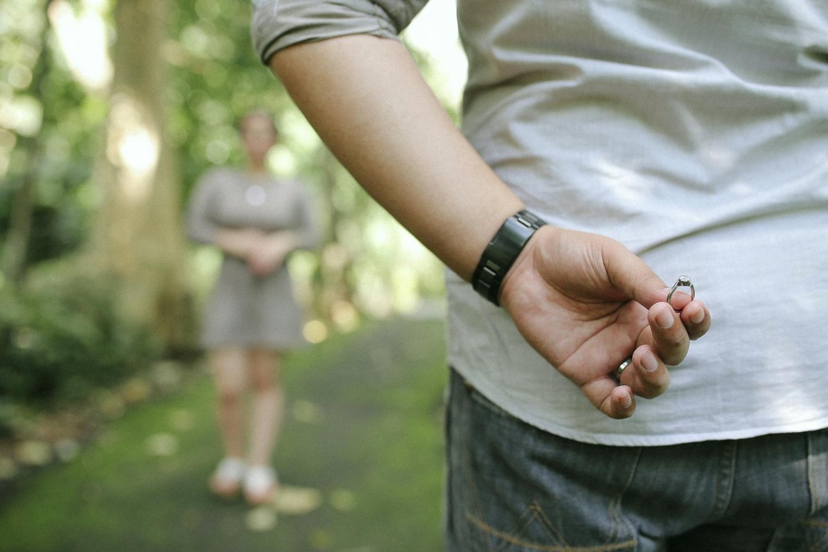 A man hides an engagement ring behind his back, preparing to propose in a picturesque park setting.
