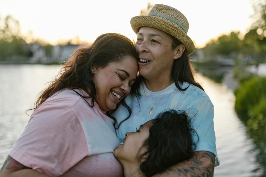 Happy same-sex family sharing an emotional hug outdoors near a serene lake at sunset.