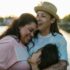 Happy same-sex family sharing an emotional hug outdoors near a serene lake at sunset.