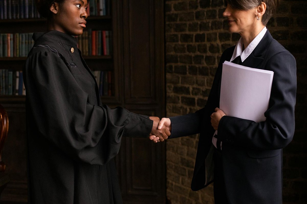 A lawyer in formal attire shakes hands with a client in a book-lined office.