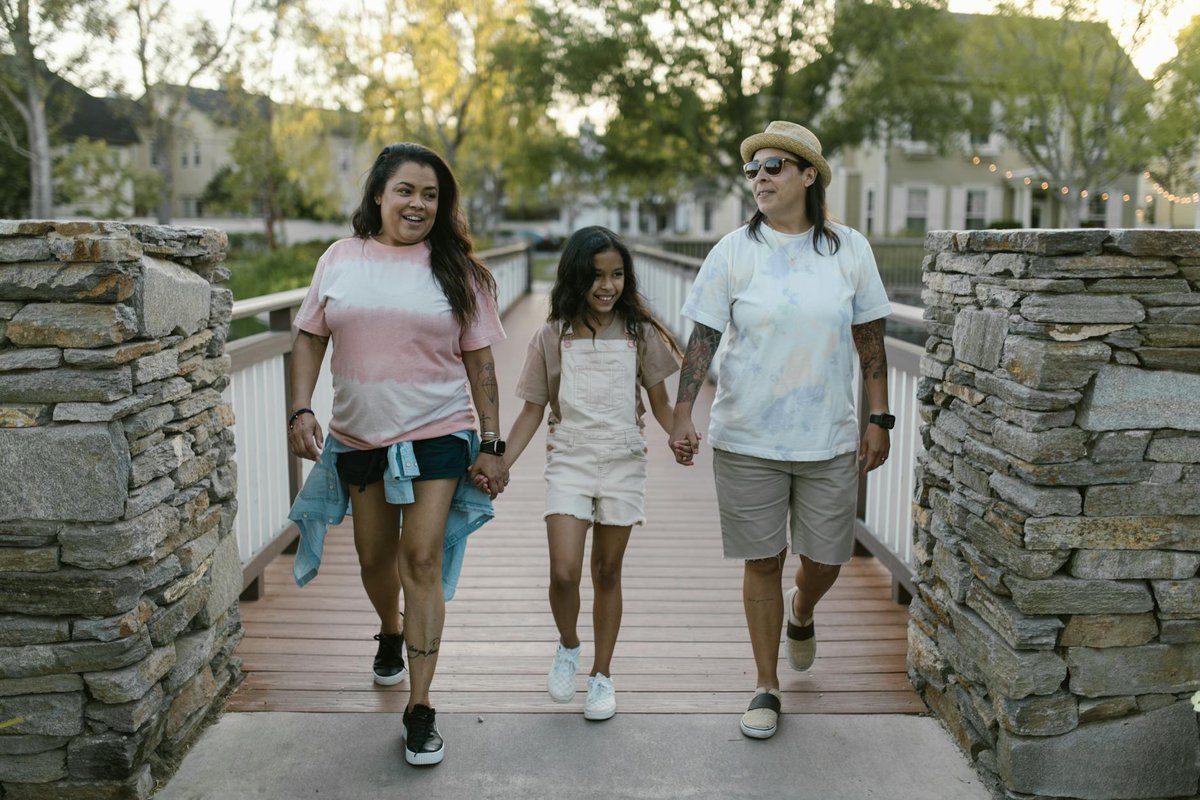A lesbian couple and their daughter enjoy a sunny day on a bridge, holding hands and smiling.