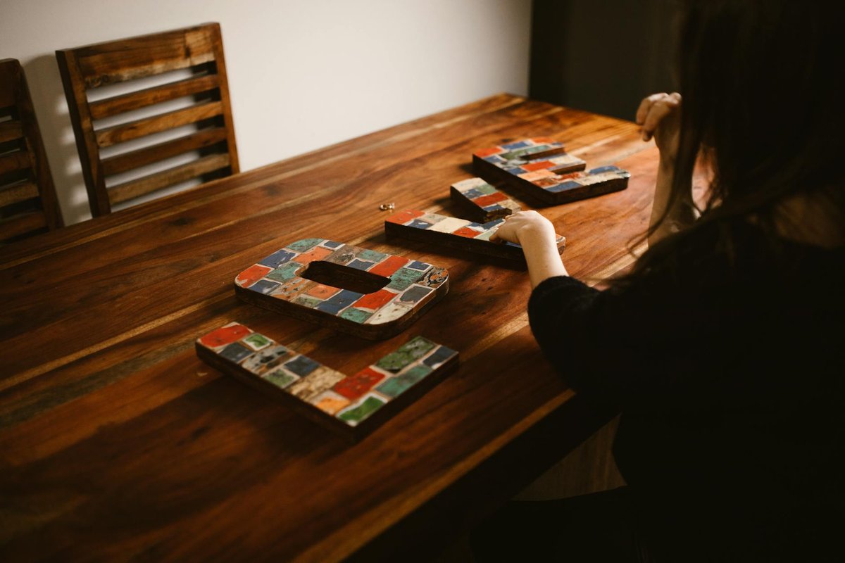 A woman arranging colorful mosaic letters on a wooden table indoors, creating art.