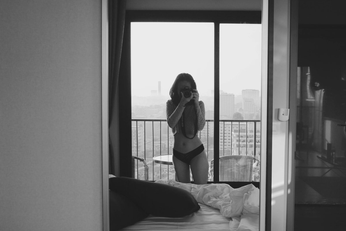 A black and white photo depicting a woman taking a selfie in a mirror inside a room with a city view.