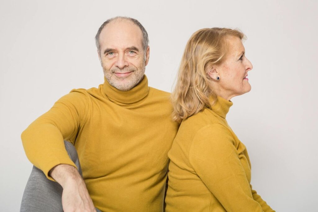 Elderly couple wearing yellow sweaters, relaxing and smiling indoors, displaying warmth and togetherness.