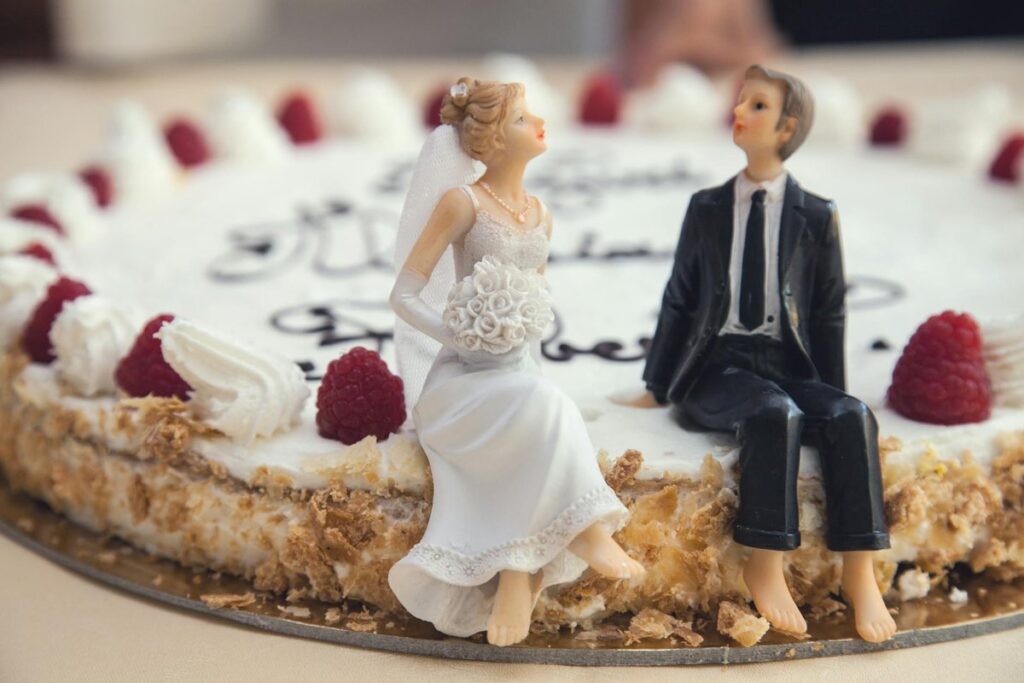 Close-up of a wedding cake with bride and groom figurines, raspberries, and whipped cream.