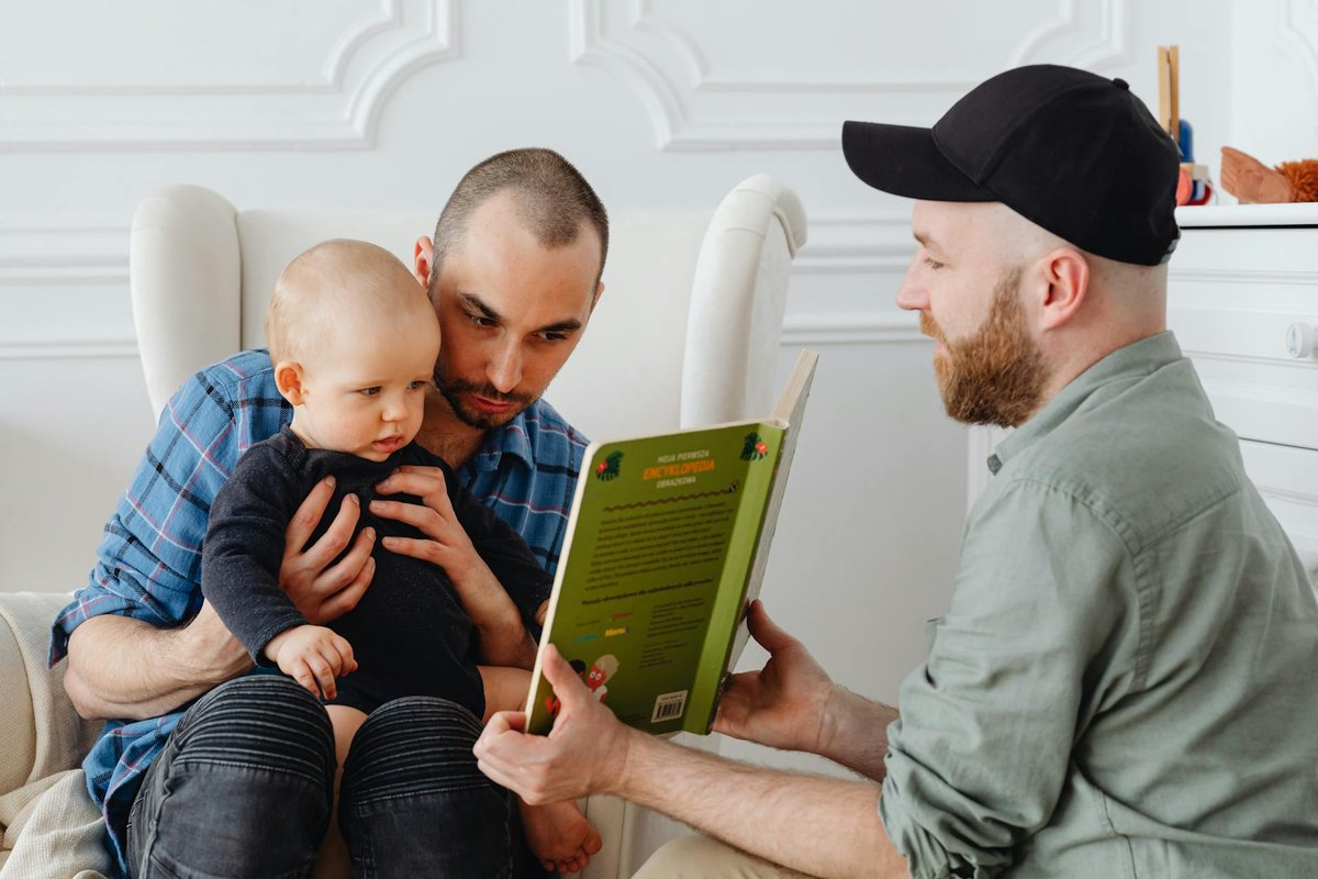 Two fathers reading a book to their toddler at home.